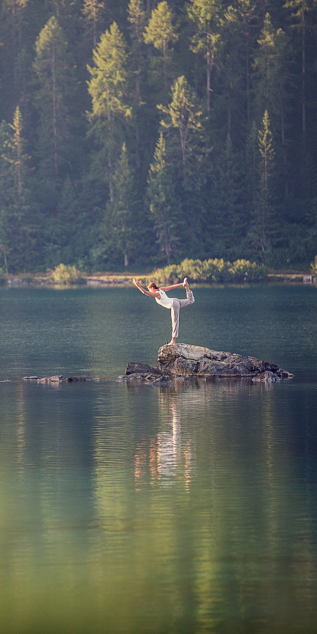 Eine Frau steht bei einer Yogaübung einbeinig auf einem grossen Stein in einem See im Oberengadin