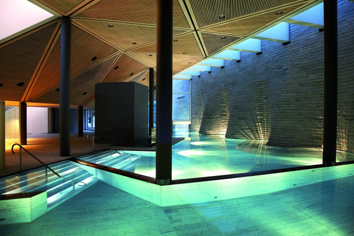 Spa-Bereich im Tschuggen Arosa Innenaufnahme eines modernen Spa-Bereichs mit beleuchtetem Pool, Treppenstufen ins Wasser und einer hohen Steinwand.