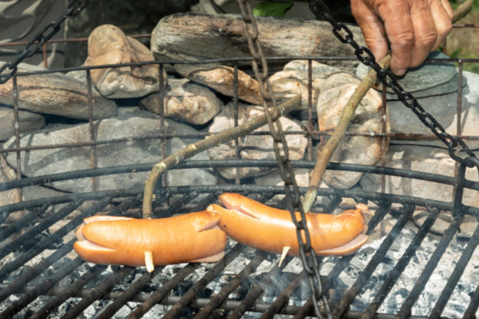 Das Foto zeigt eine Feuerstelle und zwei Cervelats die am braten sind Das Foto zeigt eine Feuerstelle und zwei Cervelats die am braten sind