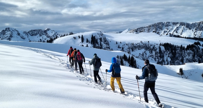 Gruppe auf Schneeschuhen folgt einer Spur über verschneite Hänge, dahinter Bergkämme und Wolkenhimmel	