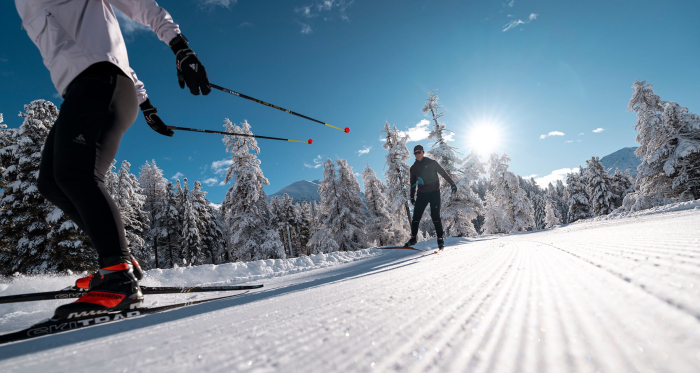 Zwei Personen beim Skating-Langlauf auf einer frisch präparierten Loipe durch einen tief verschneiten Winterwald bei Sonne