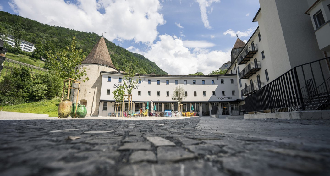 Bogentrakt Hostel Chur (Foto: © Marco Hartmann) Aussenansicht des Bogentrakt Hostels im Sennhof Chur mit historischem Rundturm heller Fassade und bunter Terrasse vor grünem Berghintergrund
