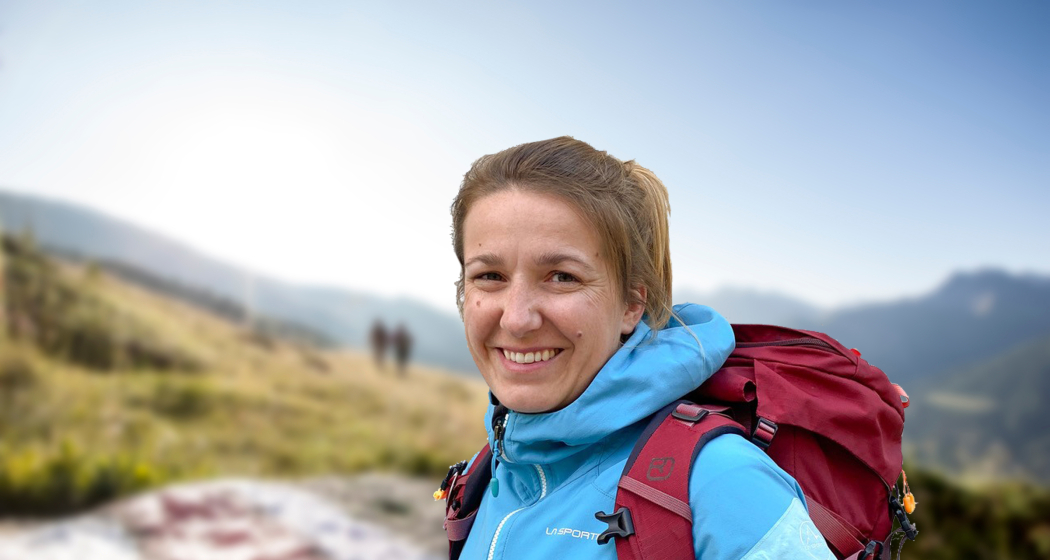 Wanderleiterin Elisabeth Hunziker begleitet Gäste auf genussvollen Wander- und Schneeschuhtouren zu den Naturperlen Graubündens Wanderleiterin Elisabeth Hunziker steht mit rotem Rucksack und blauer Jacke lächelnd auf einem Bergweg vor verschwommenem Alpenhintergrund