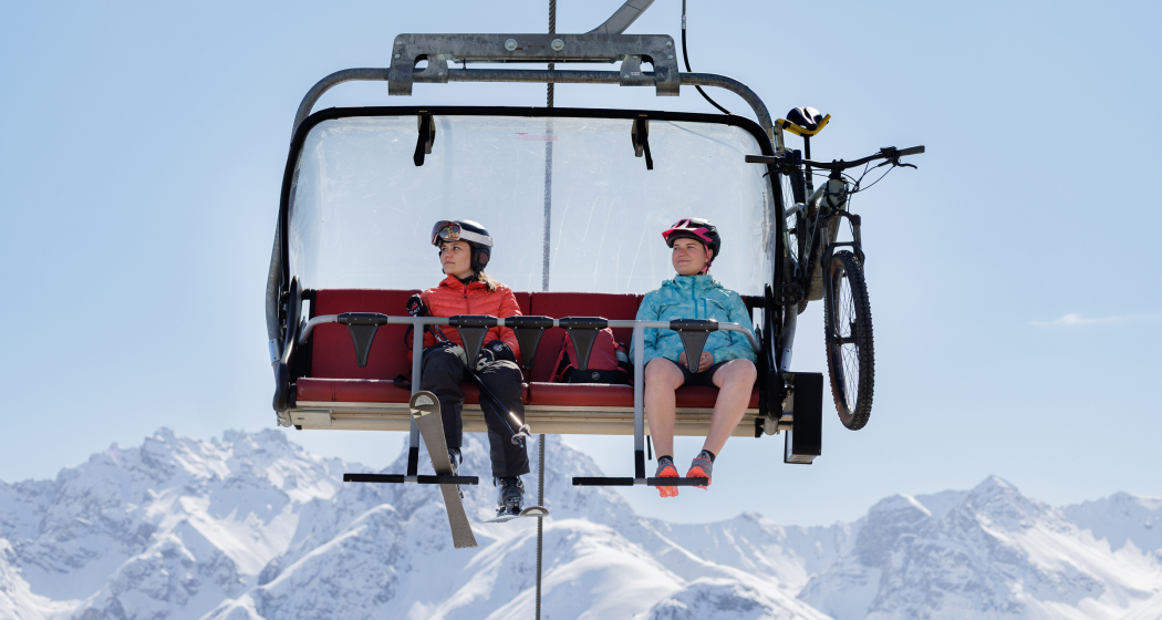 Eine Skifahrerin mit Skiausrüstung und eine Mountainbikerin mit Velo und Helm nebeneinander auf einem Sessellift vor schneebedeckten Bergen