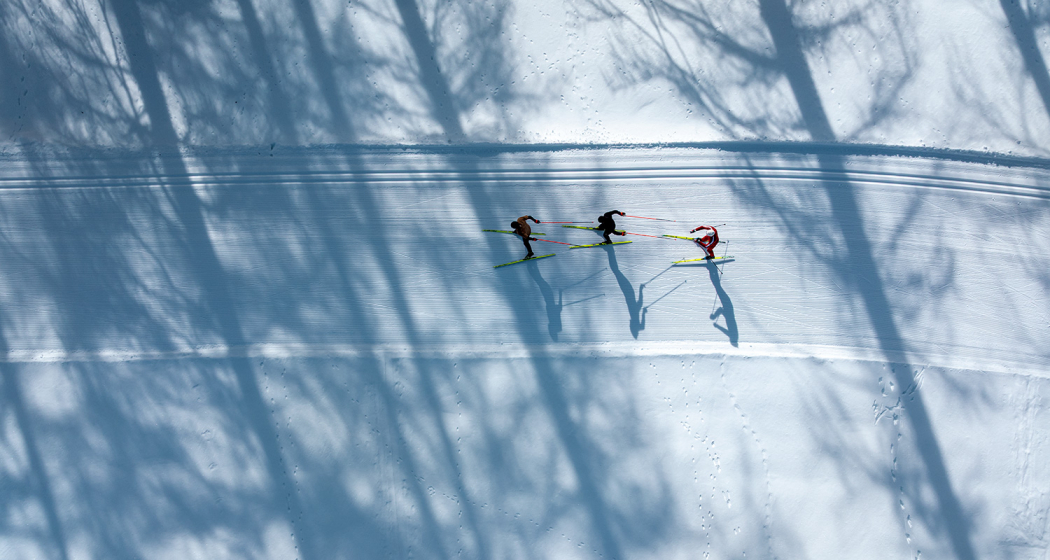 Luftaufnahme von drei Personen beim Langlaufen auf einer Loipe mit langen Baumschatten im Schnee