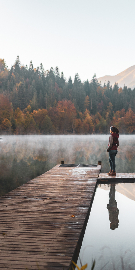 Eine Frau steht auf einem Holzsteg und blickt bei herbstlicher Stimmung auf den Crestasee