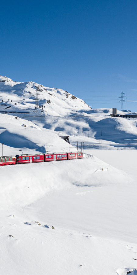 The Bernina Express travels along Lago Bianco, surrounded by snow-capped mountains and a picturesque winter landscape