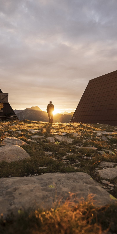Due bivacchi a forma di Toblerone sulla terrazza del Pian Grand, con un escursionista all’alba e le montagne sullo sfondo