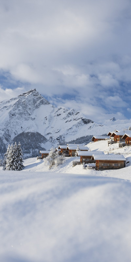 Verschneite Holzchalets des Maiensässresorts Aclas Heinzenberg vor winterlicher Bergkulisse
