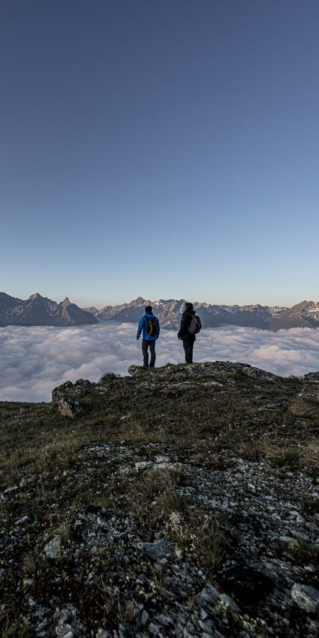 Two hikers standing on Mot da l’Hom above a sea of clouds with a view of the mountains in the Lower Engadine