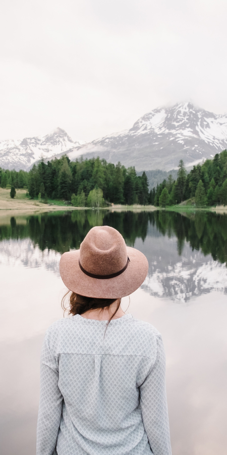 Eine Frau mit Hut blickt auf den Lej da Staz, in dem sich die Berge spiegeln