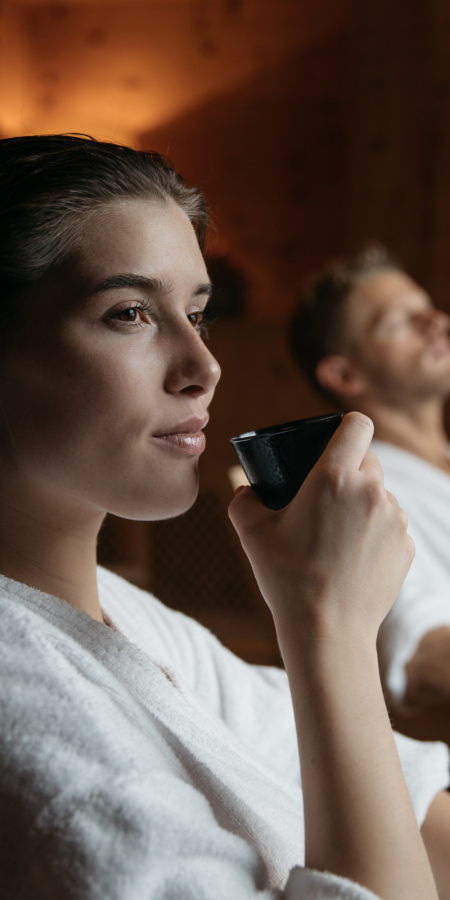 Femme en peignoir tenant une tasse dans l’espace de repos du Mineralbad Samedan, avec un homme qui se détend en arrière-plan