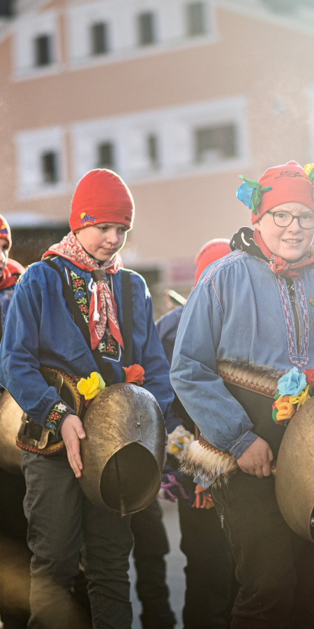 Des enfants et des jeunes en chemises bleues et bonnets rouges portent de grosses cloches à travers un village