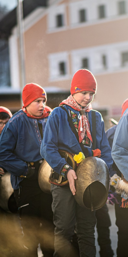 Des enfants et des jeunes en chemises bleues et bonnets rouges portent de grosses cloches à travers un village