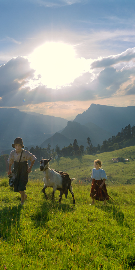 Deux enfants en tenue traditionnelle marchent avec une chèvre dans un alpage verdoyant et ensoleillé à Maienfeld, avec la silhouette brumeuse de la vallée du Rhin en arrière-plan