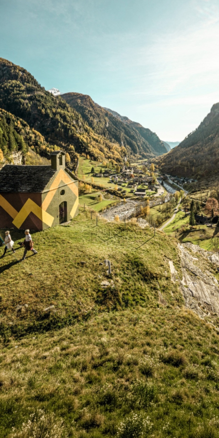 Deux randonneurs passent devant une chapelle historique ornée de peintures murales géométriques marron et jaune, située sur une colline dominant la vallée verdoyante de Calanca près de Rossa