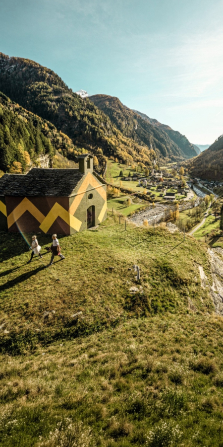Deux randonneurs passent devant une chapelle historique ornée de peintures murales géométriques marron et jaune, située sur une colline dominant la vallée verdoyante de Calanca près de Rossa