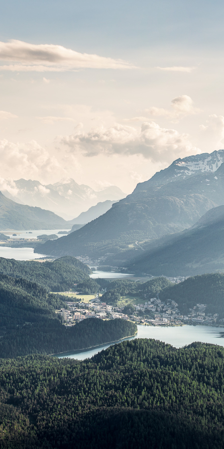 Blick von oben auf die im Tal liegende Oberengadiner Seenplatte um St. Moritz, gerahmt von bewaldeten Hängen und schneebedeckten Berggipfeln unter leicht bewölktem Himmel