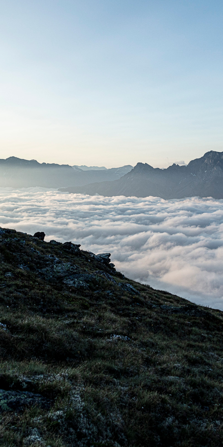 Zwei Personen stehen mit Rucksäcken auf einer Grasflanke hoch über einem dichten Nebelmeer und blicken auf eine ferne Berggipfelkette