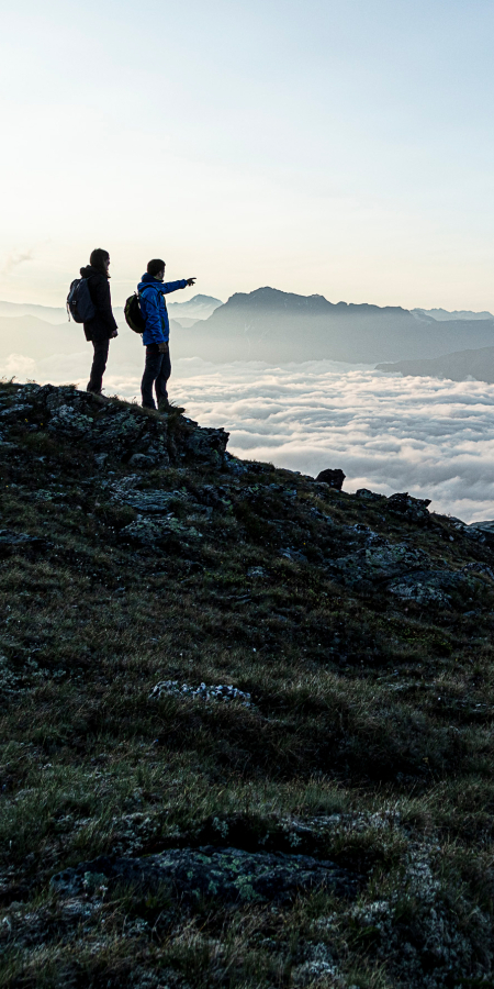 Zwei Personen stehen mit Rucksäcken auf einer Grasflanke hoch über einem dichten Nebelmeer und blicken auf eine ferne Berggipfelkette
