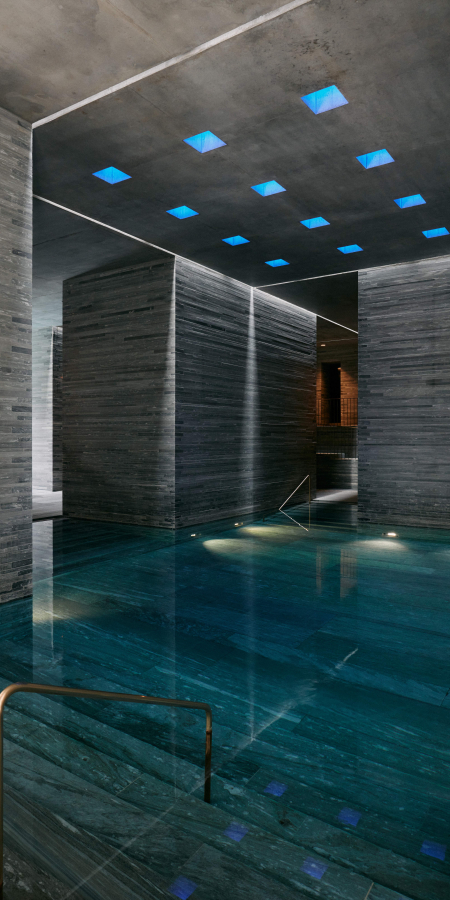 Innenansicht der Therme Vals aus grauem Valser Quarzit mit einem blau schimmernden Thermalbecken und sanft herabhängenden Hängelampen