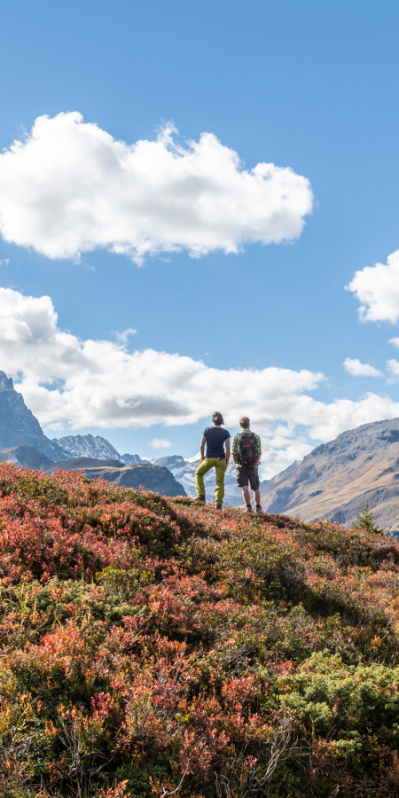 Two hikers stand on a reddish-brown autumn meadow on the Alp Flix, looking out at the surrounding rocky mountain peaks of the Val Surses