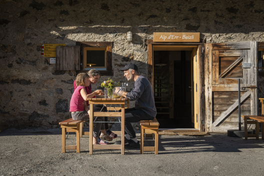 Alp d'Err im Val Surses (Foto: © Tom Malecha / Schweiz Tourismus) Eine Frau, ein Mann und ein Mädchen sitzen vor der Alp d'Err und essen zu Abend.