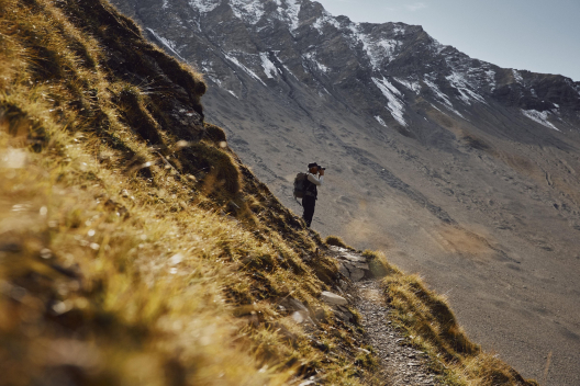 Le garde Domenic Godly en mission d’observation dans la vallée de Trupchun, au Parc national suisse (Photo : © Suisse Tourisme / Colin Frei) Le garde Domenic Godly se tient avec des jumelles sur un sentier de montagne dans la vallée de Trupchun, face à un paysage rocailleux