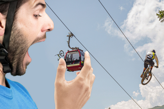 Ein übergrosser Mountainbiker beisst in die Gondel einer Bergbahn