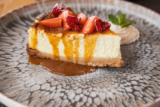 Kuchen mit Karamellsosse und frischen Erdbeeren liegt auf einem dekorativen Teller. Daneben befindet sich ein Klecks Sahne, garniert mit einem Minzblaettchen.
