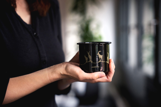Person hält eine schwarze Emaille-Tasse mit goldenen Steinbockmotiven und graubünden-Logo in den Händen