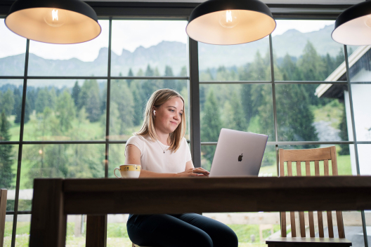 Workation in Graubünden: Junge Frau arbeitet am Laptop vor einem grossen Fenster mit Blick in die Berge