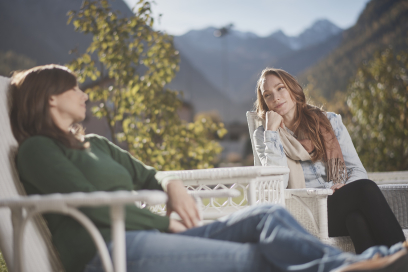 Achtsamkeit in der Natur – gemeinsames Innehalten mit Blick auf die Berge (Foto: © Graubünden Ferien / Samuel Trümpy) Zwei Frauen sitzen entspannt auf weissen Terrassenstühlen und unterhalten sich, im Hintergrund die Berge bei Bergün