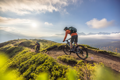 Goldenes Licht, grüne Hügel und pure Trailfreude in Disentis Sedrun (Foto: © WOM Medien) Zwei Mountainbiker fahren bei Sonnenaufgang auf einem flowigen Singletrail durch die grüne Berglandschaft in der Ferienregion Disentis Sedrun