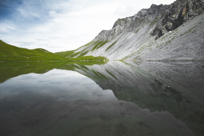 Ein ruhiger Bergsee spiegelt die umliegenden grauen Felsen und gruenen Wiesen