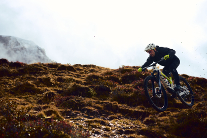 Ein Mountainbiker mit Helm und Handschuhen fährt eine steile, moosbewachsene Bergwiese hinunter, während im Hintergrund Wolken und Berge sichtbar sind.