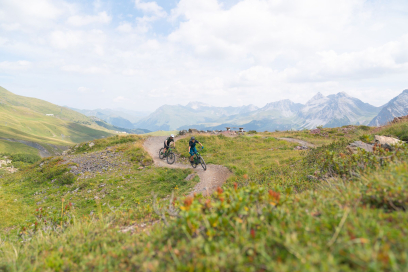 Zwei Mountainbiker fahren auf einem flowigen Singletrail durch eine grüne Alpenlandschaft mit weitem Blick auf umliegende Berge und Täler.