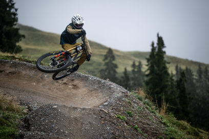 in Mountainbiker mit Helm und Schutzkleidung fährt in Schräglage über eine kurvige Schotterstrecke im Gebirge, während im Hintergrund grüne Hügel und Bäume zu sehen sind.