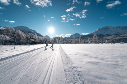 Zwei Langläufer auf einer verschneiten Loipe im Engadin, umgeben von schneebedeckten Bäumen und den Bergen im Hintergrund, bei sonnigem Wetter.