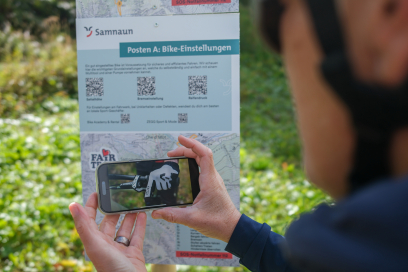 Mountainbike Skill Loop in Samnaun – Übungen und Tipps direkt im Gelände (Foto: © Franz Balmer) Person hält ein Smartphone vor eine Infotafel des Mountainbike Skill Loops in Samnaun, auf dem ein Video zur Bike-Einstellung angezeigt wird