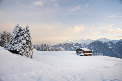 Verschneite Berglandschaft mit schneebedeckten Tannen im Vordergrund, einer einzelnen Berghütte rechts und einer Reihe hoher, winterlicher Berggipfel im Hintergrund unter einem sanft bewölkten Himmel.