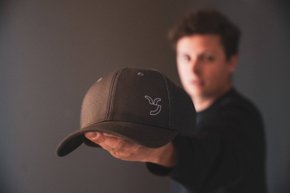 Eine Person hält eine dunkelgraue Baseballcap mit weissem Capricorn-Logo in die Kamera; der Hintergrund und die Person sind unscharf gehalten.