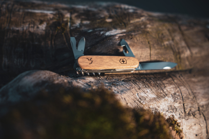 Victorinox Taschenmesser Huntsman Wood aus Schweizer Nussbaumholz mit ausgeklappten Werkzeugen und eingraviertem Steinbock-Logo auf einem verwitterten Baumstamm