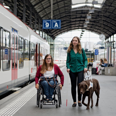 L'accès surbaissé de nombreux trains facilite l'embarquement et le débarquement dans de nombreuses gares (Photo : © SBB CFF FFS) Une femme en fauteuil roulant et une femme avec un chien guide d'aveugle sur un quai de gare