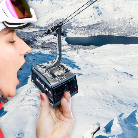 Eine übergrosse Skifahrerin in voller Montur beisst spielerisch in eine Gondel einer Bergbahn