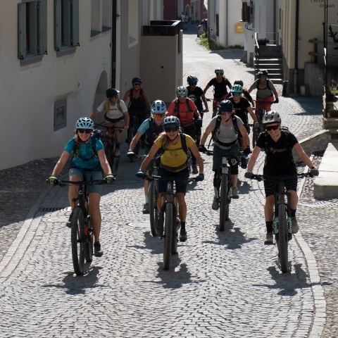 Eine Gruppe von etwa 15 Radfahrerinnen fährt bei sonnigem Wetter durch eine enge, gepflasterte Gasse in einem historischen Dorf