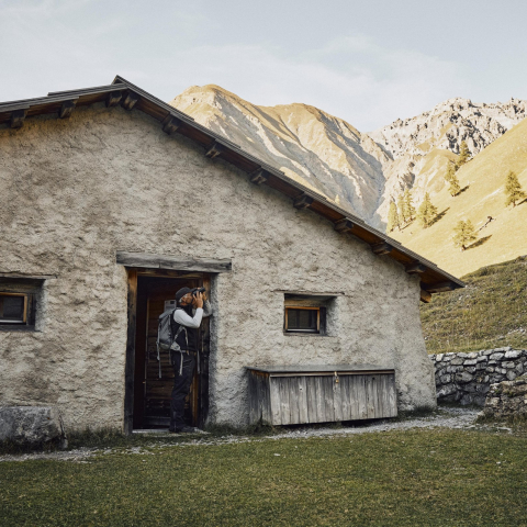 Le garde du Parc national Domenic Godly devant la cabane du parc dans la vallée de Trupchun – au cœur sauvage du Parc national suisse (Photo : © Suisse Tourisme / Colin Frei) Garde du parc national avec des jumelles devant une cabane dans la vallée de Trupchun, au cœur du Parc national suisse, entouré de montagnes alpines