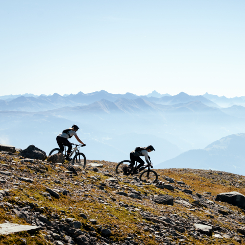 Trailvergnügen mit Aussicht (Foto: © Schweiz Tourismus) Zwei Mountainbikerinnen fahren über einen felsigen Bergtrail mit spektakulärer Aussicht auf ein weites Alpenpanorama