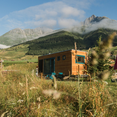 Kleines rechteckiges Tiny Home auf dem Naturcampingplatz Viva in Rueras mit blauen Fenstern und Türen inmitten einer Wiese, vor grünen Bergen mit Wolken und Felsen im Hintergrund
