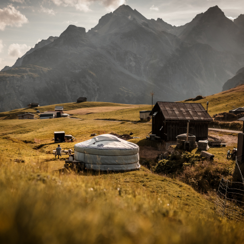 Berglandschaft mit traditionellen Holzhäusern und einer weissen Jurte auf einer Alp, umgeben von sanften Wiesen und hohen Gipfeln im Hintergrund.